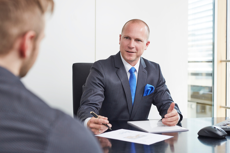 Ein Mann im Anzug führt ein Gespräch mit einem Kollegen an einem Schreibtisch.